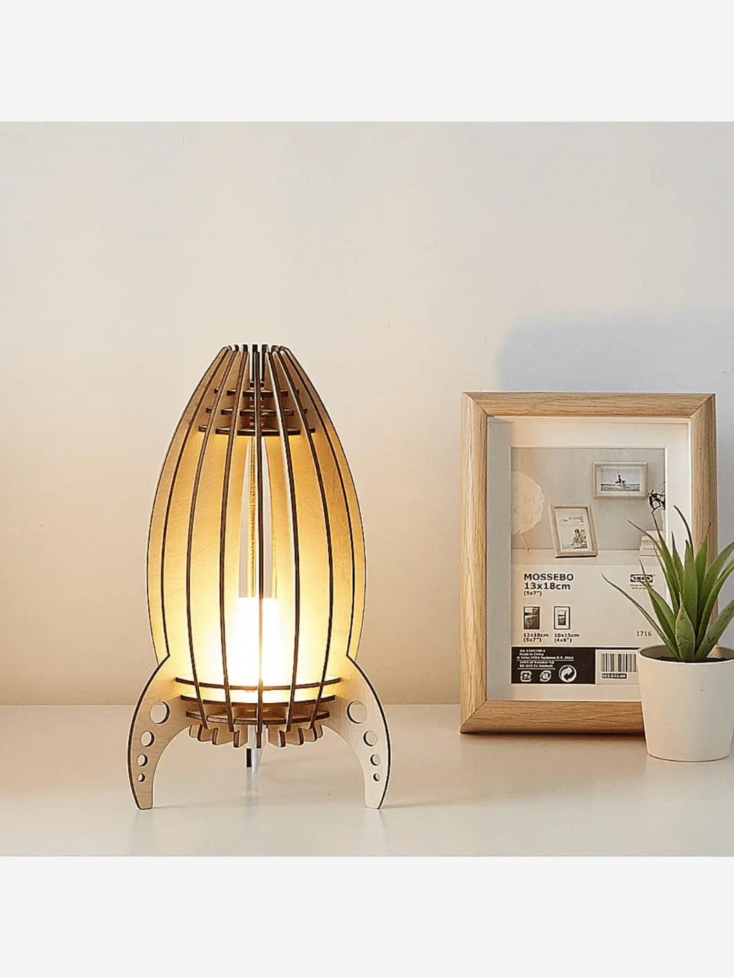 lampe de chevet garcon fusee en bois allumee et posee sur une table de chevet blanche a cote d un cadre et d une plante
