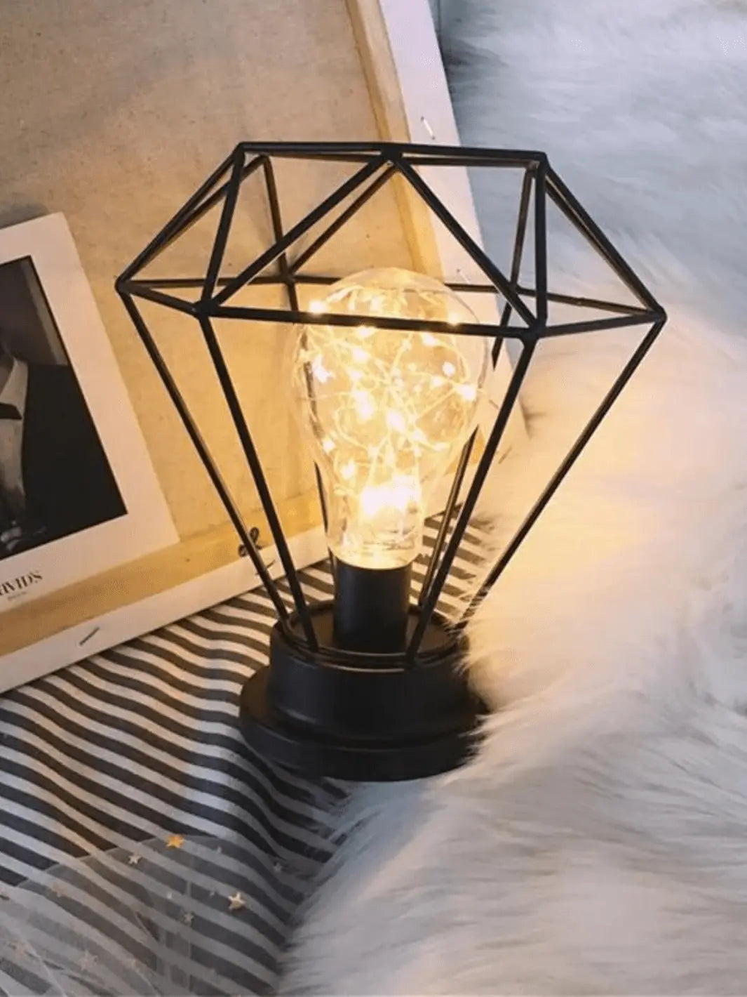 lampe de chevet industrielle en metal en forme de diamant noir pose sur un socle avec une ampoule led a l interieur allumee posee dans un decor cozy