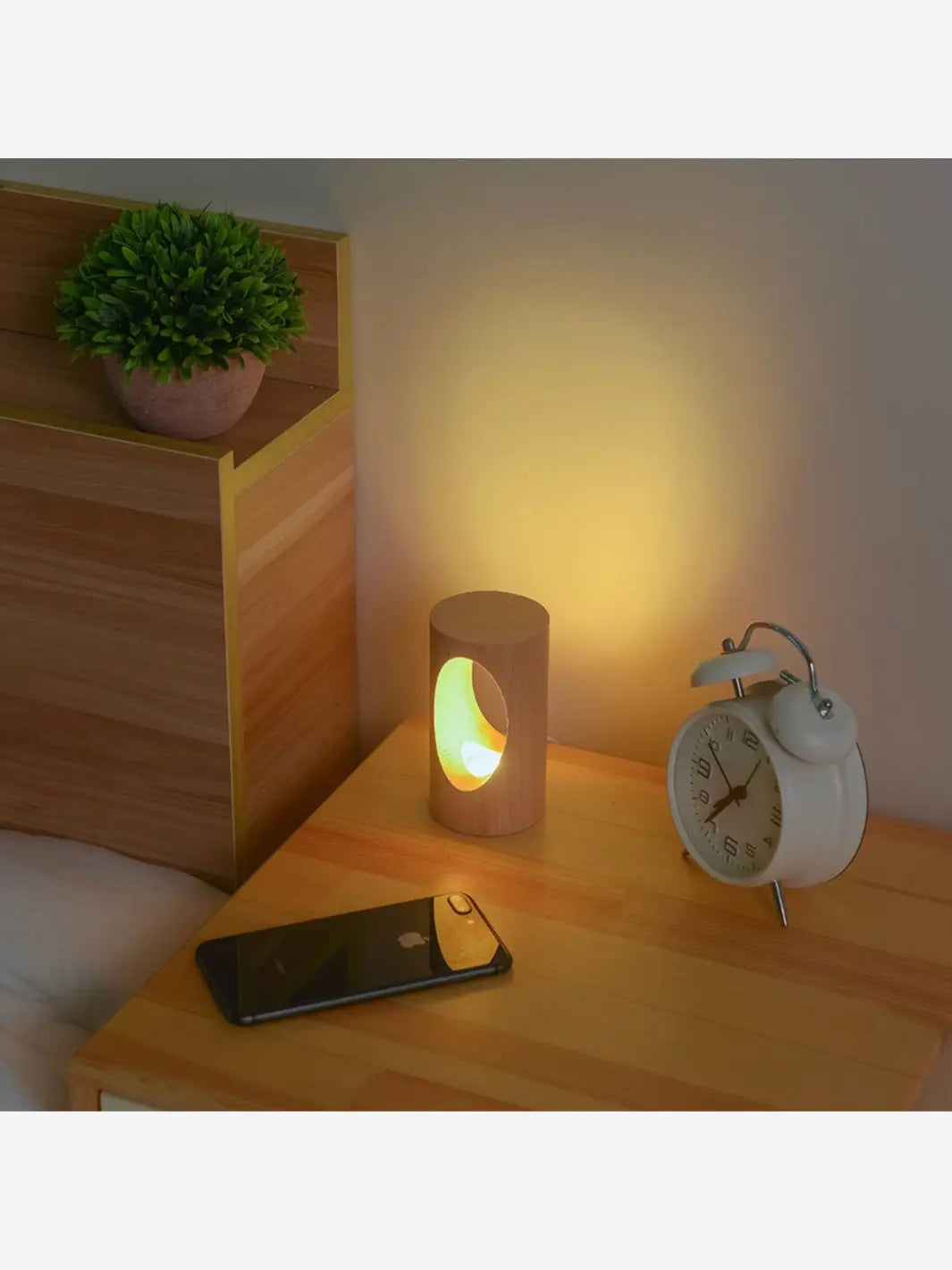 lampe de chevet scandinave en bois eclairee posee sur une table de chevet en bois avec un reveil et un smartphone